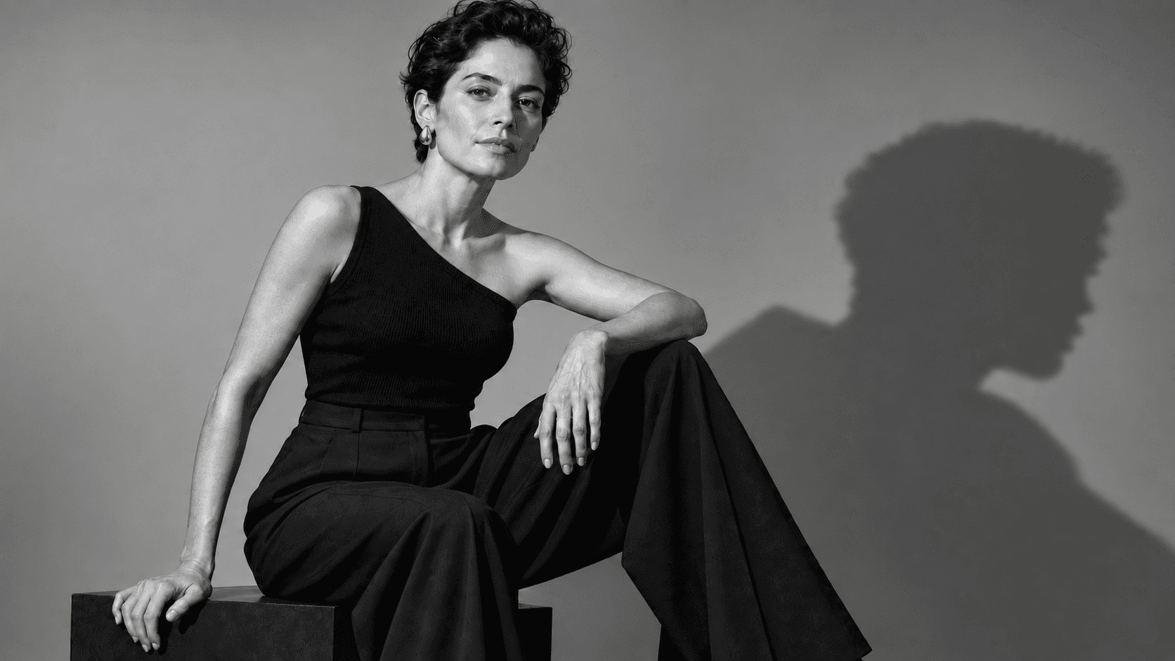 Black-and-white editorial portrait of a woman in an asymmetric black top, with her jawline visible and a profile shadow behind her.