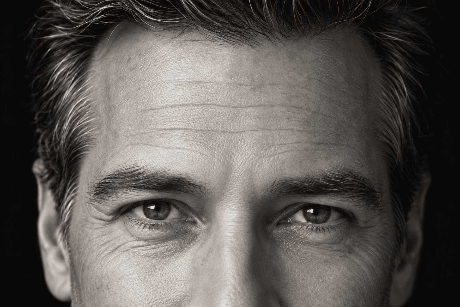 Black-and-white editorial close-up of an adult man’s forehead and eyes, showing natural skin texture and expression lines.
